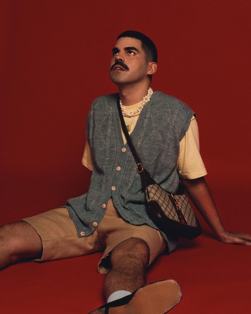 Mateus Carrilho, vestido de colete, bermuda, camiseta e bolsa de lado, em um fundo vermelho, olhando para cima.