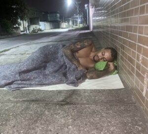 Meme mostrando uma pessoa dormindo na rua esperando a sua vez de ser ajudado pela mulher do personal trainer