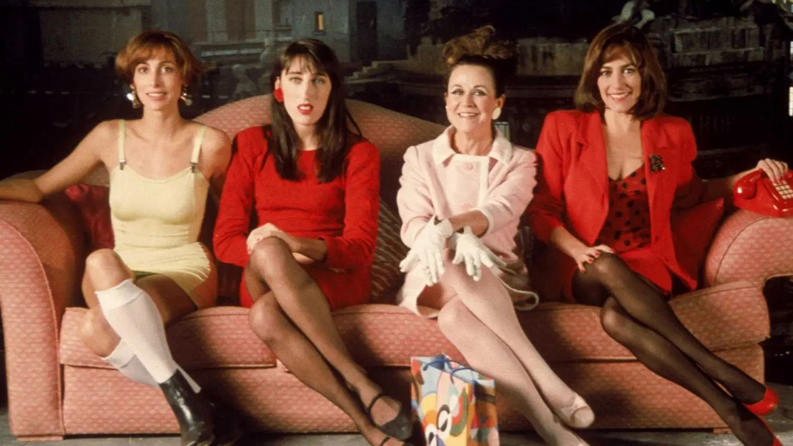 A imagem mostra as atrizes principais do filme "Mulheres à beira de um ataque de nervos", caracterizadas como suas personagens. Da direita para a esquerda aparecem María Barranco como Pepa; Rossy de Palma, como Marisa; Julieta Serrano, como Lucía e Carmen Maura como Pepa. Todas estão sentadas, da esquerda para a direita, nessa ordem, num sofá de três lugares, rosa. María se veste de amarelo claro, Julieta de rosa bebê, enquanto Rossy e Carmne vestem-se de vermelho intenso, cor marcante, cujo uso é uma das principais características da obra visual de Almodóvar.